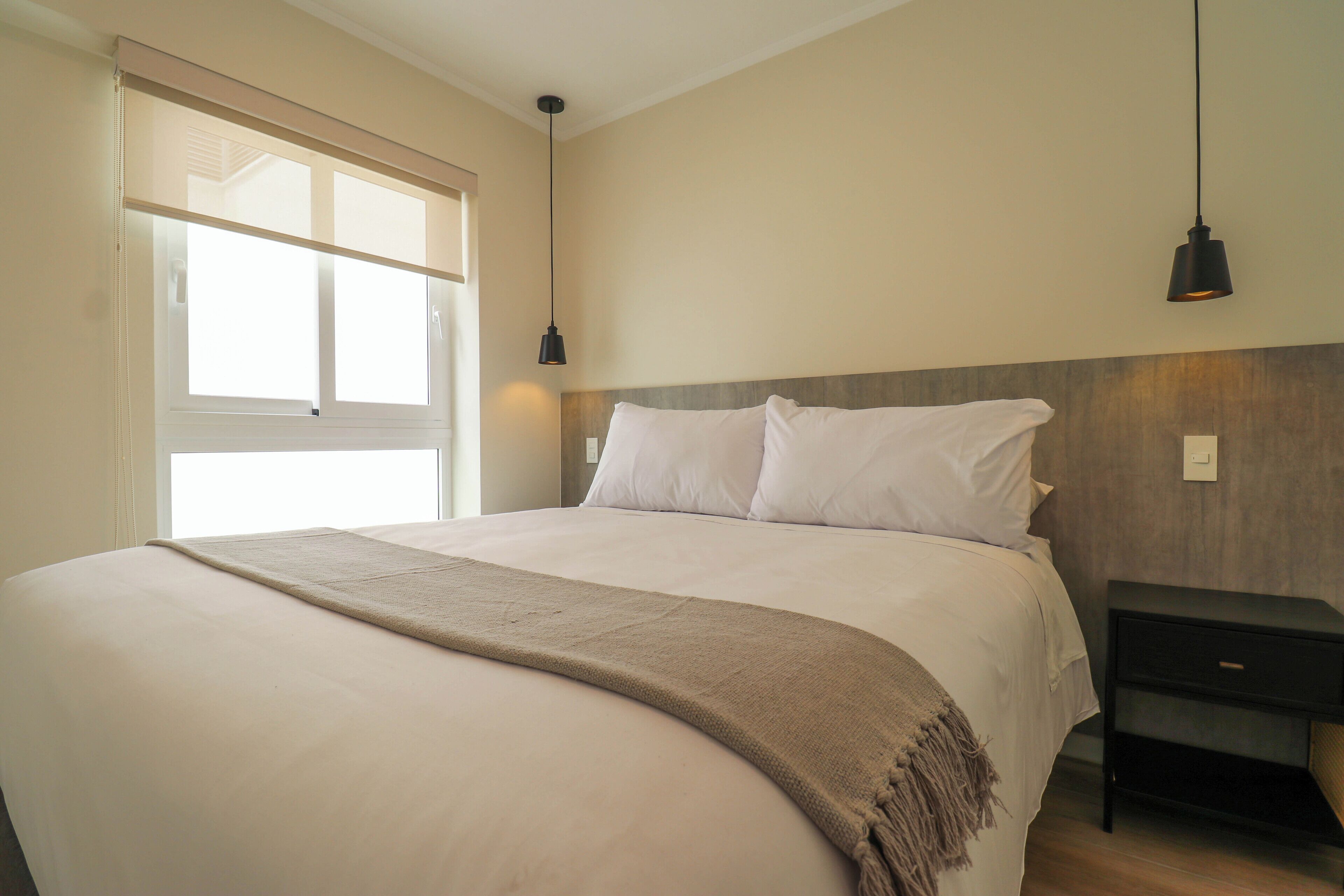 Un letto rifatto con una coperta beige, una testiera in legno e un comodino nero con un cassetto.