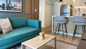 A modern living room with a teal sofa, a coffee table, and a kitchen area with a refrigerator, sink, and bar stools.