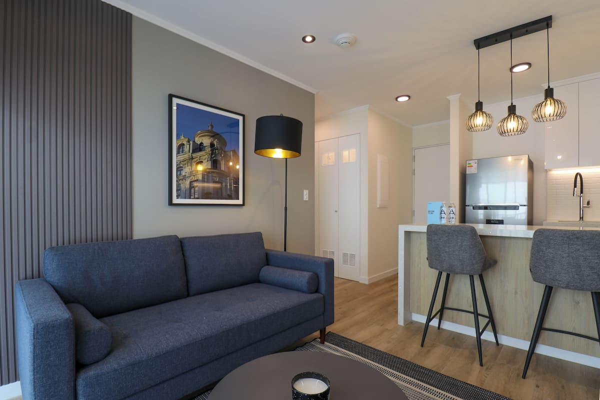 Een moderne woonkamer met een bank, salontafel en vloerlamp. Er is een keukenhoek met een koelkast en barstoelen.