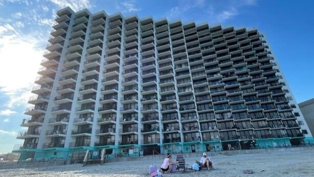 Korkea hotellirakennus, jossa on useita parvekkeita, hiekkapohjainen alakerta ja kirkkaan sininen taivas.