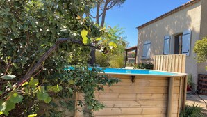 Une piscine en bois, entourée d’une terrasse en bois, située devant une maison avec un toit en tuiles et des fenêtres.