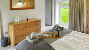 Ein Schlafzimmer mit einem hölzernen Schminktisch, einem Bett mit blumigem Überwurf, einem Tablett mit Handtüchern und einem Buch sowie Blick auf einen grünen Garten durch das Fenster.