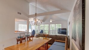 A spacious living room with a large wooden dining table, comfortable seating, a ceiling fan, and a television.