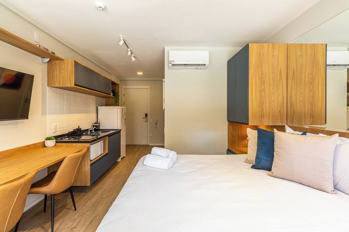 A compact hotel room with a kitchenette, a bed with pillows, a desk with a chair, and a TV mounted on the wall.