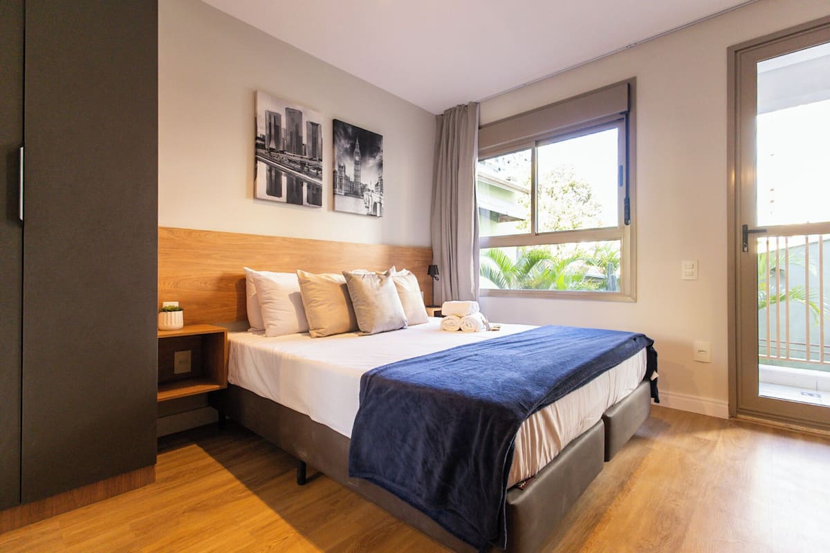 A modern hotel room with a large bed, a wooden headboard, a nightstand, and a window with a view of greenery.