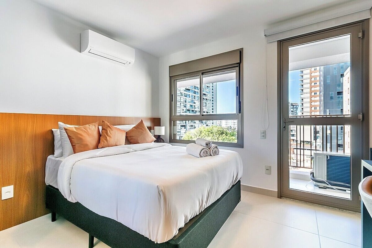A modern hotel room with a large bed, a wooden headboard, a window with a city view, and a balcony.