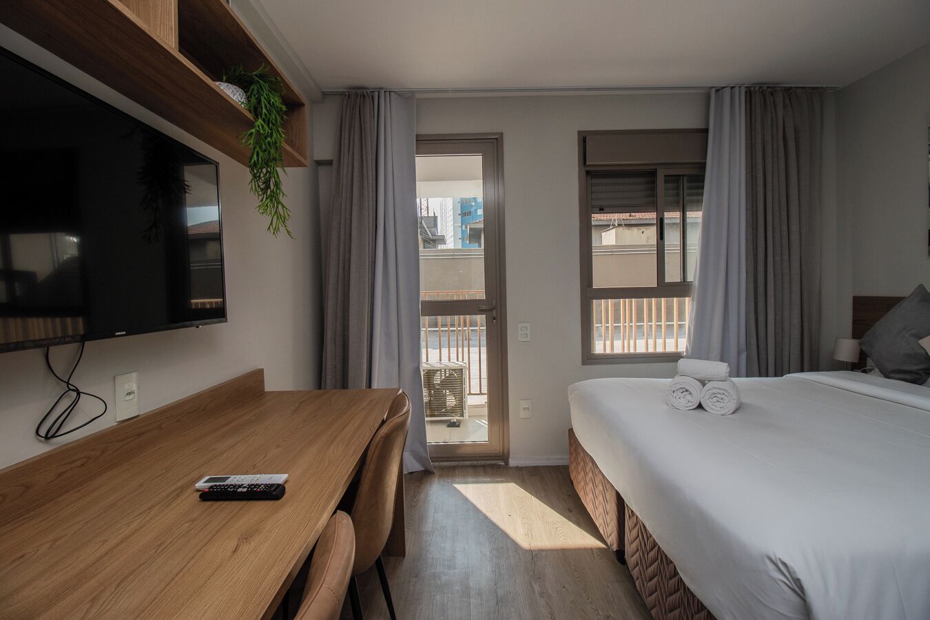A hotel room with a wooden desk, a bed with rolled towels, a TV mounted on the wall, and a balcony with curtains.