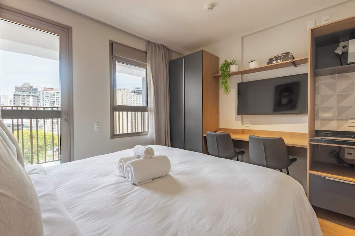 A hotel room with a large bed, a desk, a chair, a TV, and a balcony with a city view.