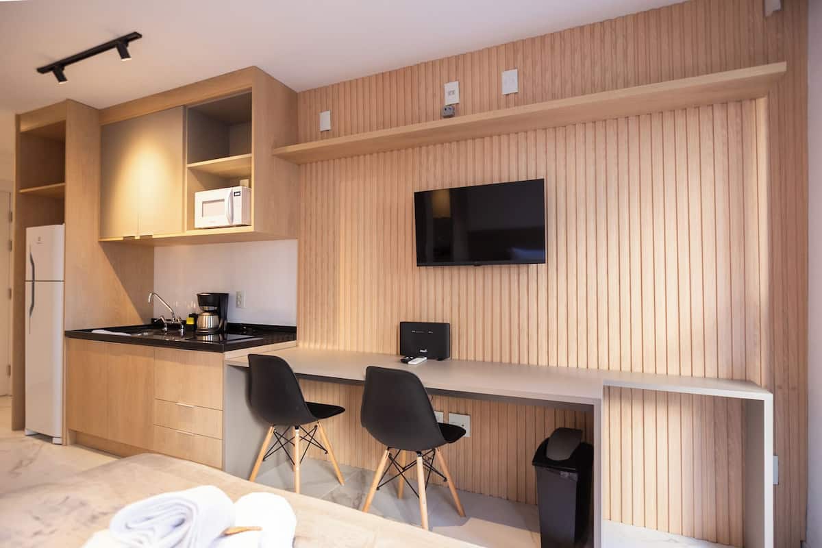 A compact kitchenette with a microwave, coffee maker, and a flat-screen TV mounted on the wall. There are two black chairs and a small trash bin.