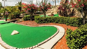 Ein Minigolfplatz mit einem grünen Putting-Bereich, umgeben von angelegten Gärten, Palmen und einem Felsen im Hintergrund.