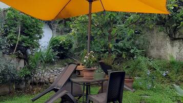 Un patio con mesa y sillas bajo un paraguas amarillo, rodeado de vegetación y un muro de piedra.