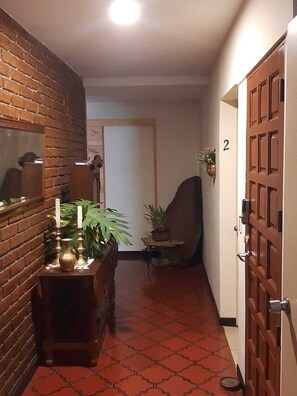 Un pasillo con pared de ladrillo, un mueble de madera con plantas, una puerta marcada con el número 2 y un suelo de baldosas.