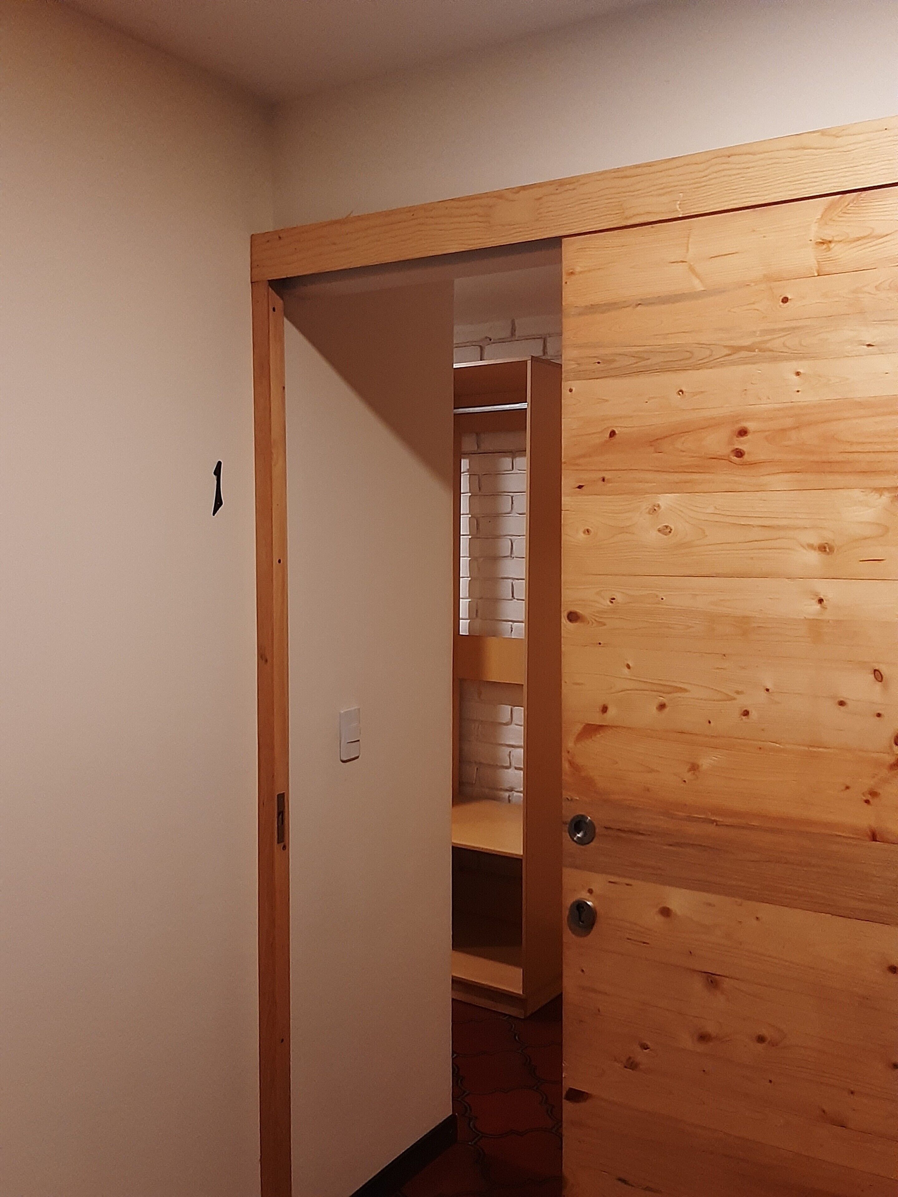 Un couloir avec une porte en bois donnant sur une pièce dont un mur est décoré d’un motif en briques et qui contient une armoire en bois.