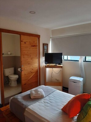 Un dormitorio con cama, televisor sobre un mueble, calefactor y ventana con persianas.