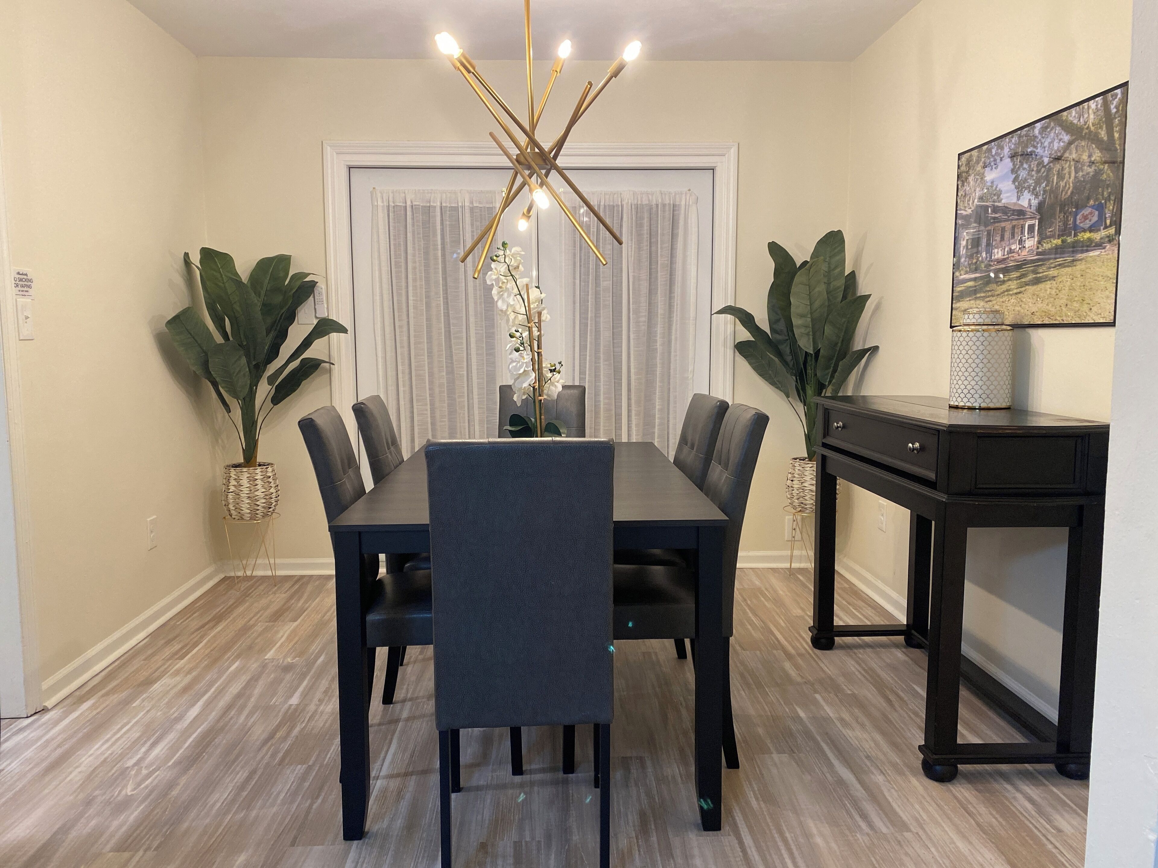 Un comedor con una mesa de madera oscura, seis sillas, una lámpara de techo moderna y plantas en macetas.