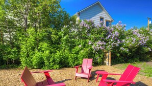 En have med røde Adirondack-stole, en træterrasse og et hus med lilla blomster i baggrunden.