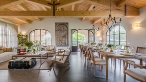 Spaziosa sala da pranzo con soffitto alto e travi in legno, ampie finestre che offrono vista sul verde, e un'accogliente area salotto con divano e tavolino.