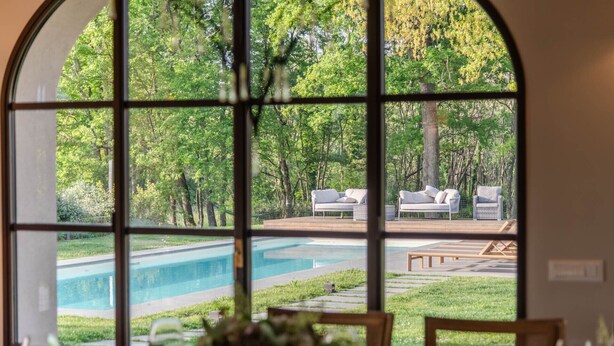 Vista attraverso un'ampia finestra su una zona piscina con posti a sedere all'aperto e un prato curato.