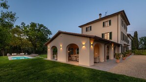 Una casa a due piani con piscina, area salotto esterna e un prato curato.