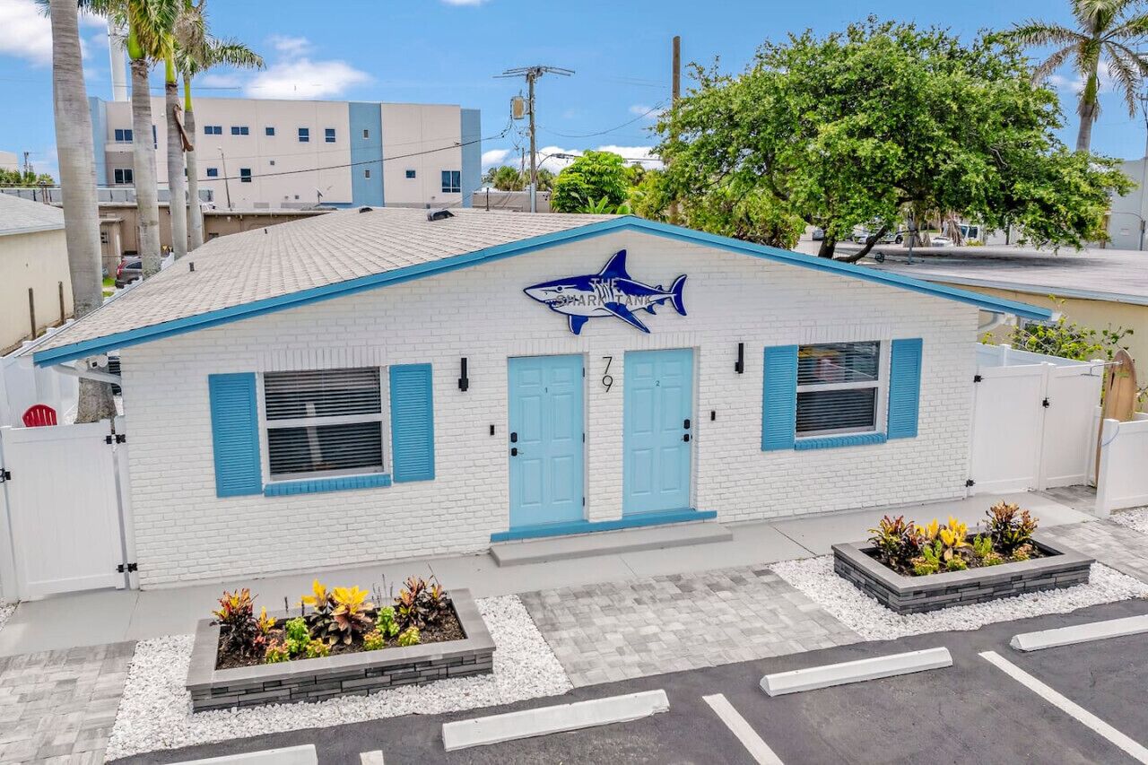 A white building with blue shutters and a shark logo, surrounded by palm trees and a parking lot.