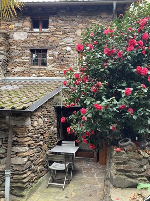 A stone building with a rustic aesthetic, a small table and chairs in the foreground, and a bush of red flowers.