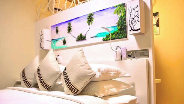 A bed with white bedding and decorative pillows, a painting of a tropical beach, and a wall-mounted power outlet. - Luna lodge  (Olhuvelifushi)