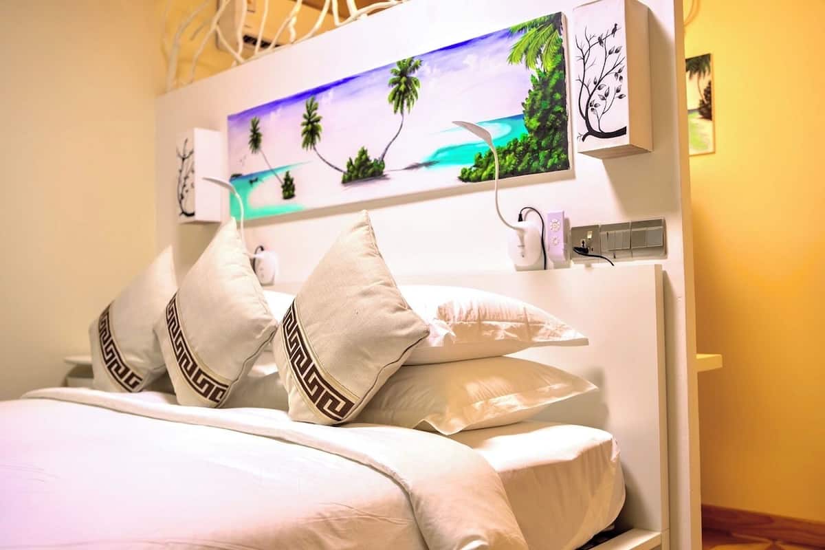 A bed with white bedding and decorative pillows, a painting of a tropical beach, and a wall-mounted power outlet.