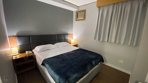 A hotel room with a bed, bedside lamps, a nightstand, a telephone, and a window with curtains. - Flat confortável próximo ao metrô (São Paulo)
