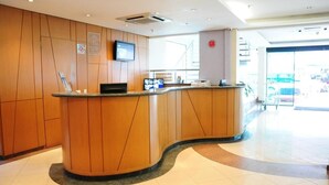 A reception area with wooden paneling, a curved reception desk, a flat-screen TV, and a glass door leading outside. - Flat confortável próximo ao metrô (São Paulo)