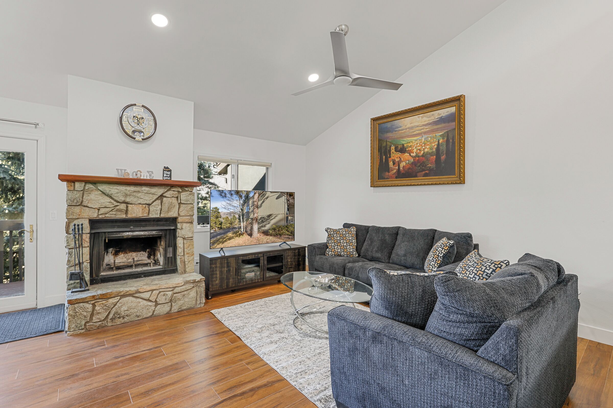 A living room with a stone fireplace, a gray sofa, a glass coffee table, and a painting on the wall.