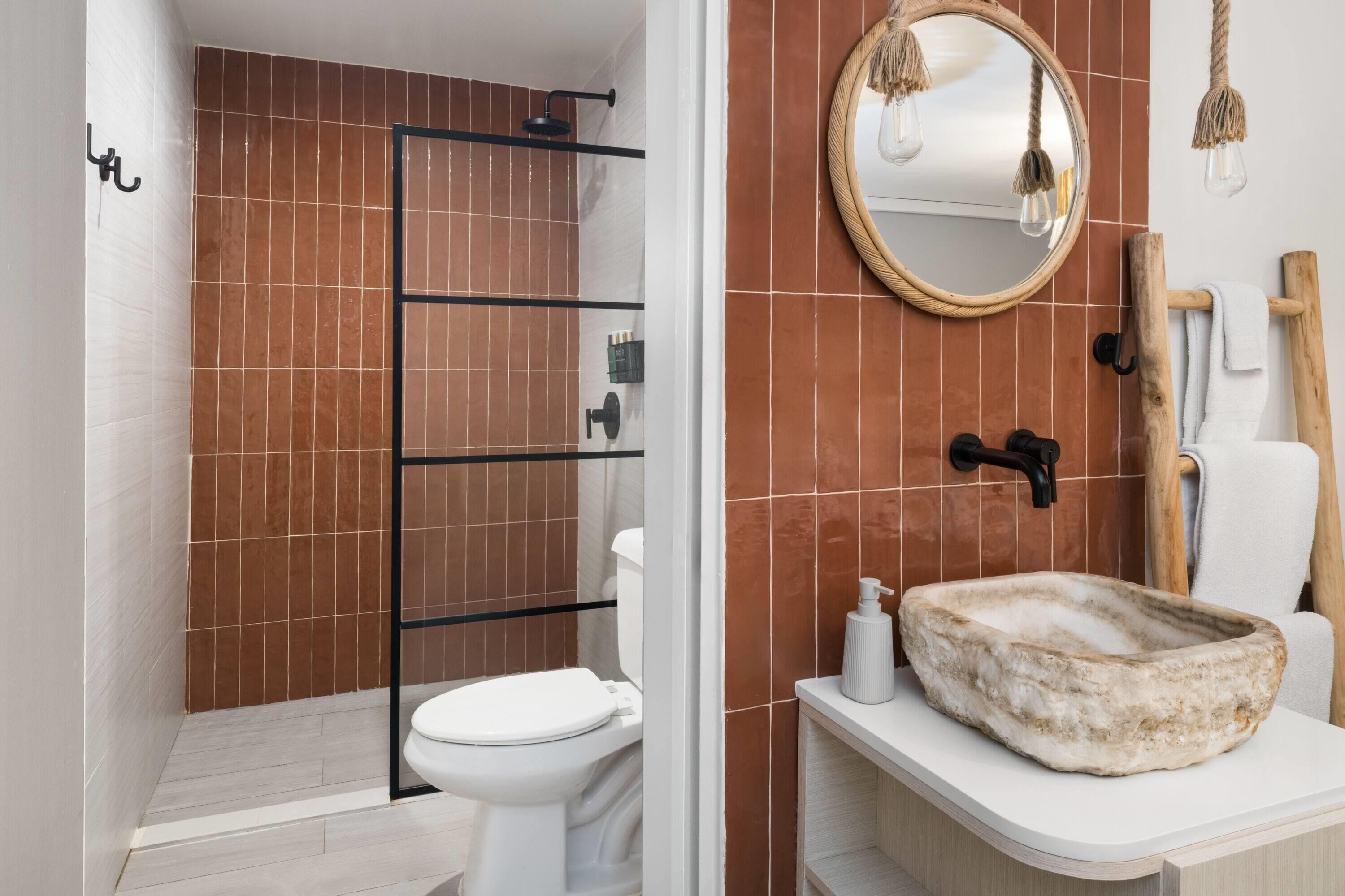 Un baño con inodoro blanco, ducha con estructura negra, espejo redondo y lavabo de piedra.