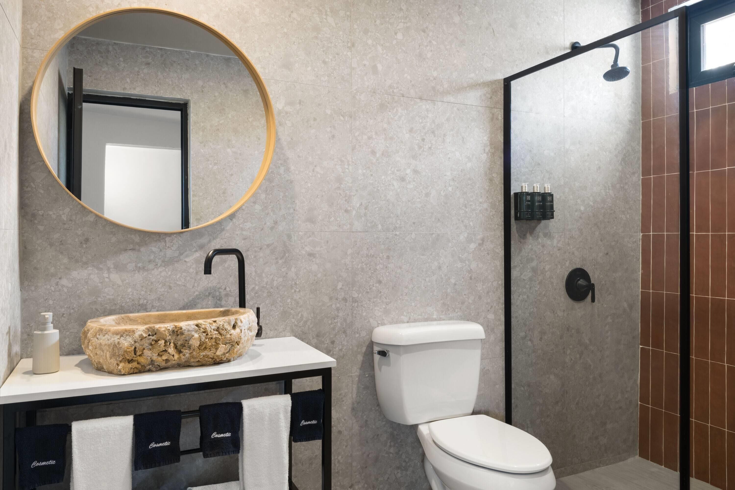A bathroom with a stone sink, a toilet, a shower, and a round mirror.