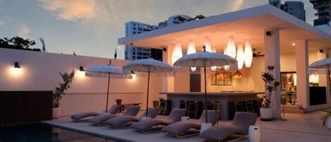 Un espace piscine extérieur moderne, doté de chaises longues et de parasols, d’un bar et d’une piscine dont l’eau reflète le ciel.