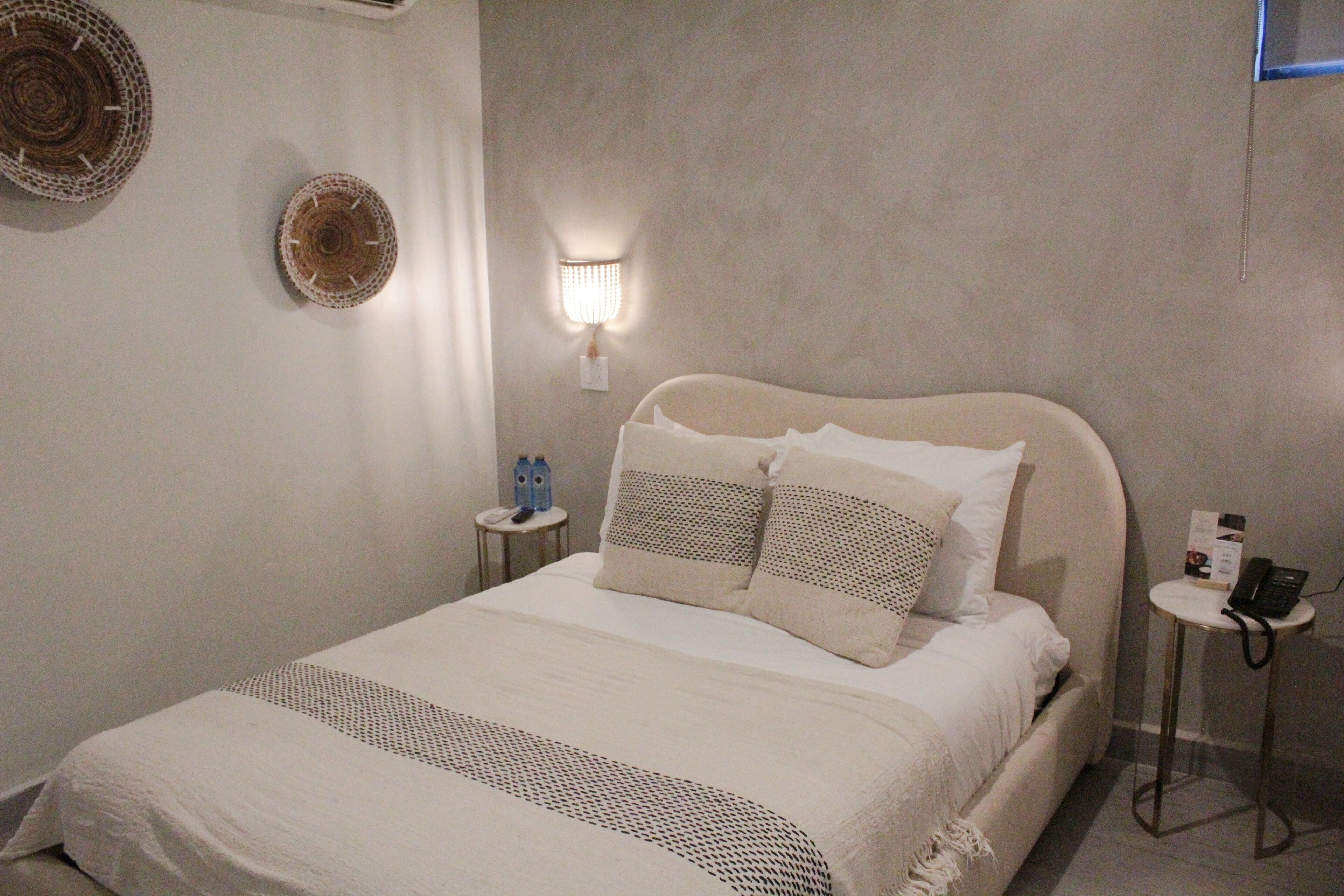 Un lit bien fait, recouvert d’une couvre-lit à motifs blancs et gris, une table de chevet avec une lampe et un luminaire fixé au mur.