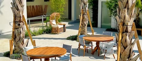 Un espace extérieur aménagé en terrasse, avec des tables rondes en bois et des chaises en osier, entouré de grands palmiers et avec un bâtiment moderne en arrière-plan.
