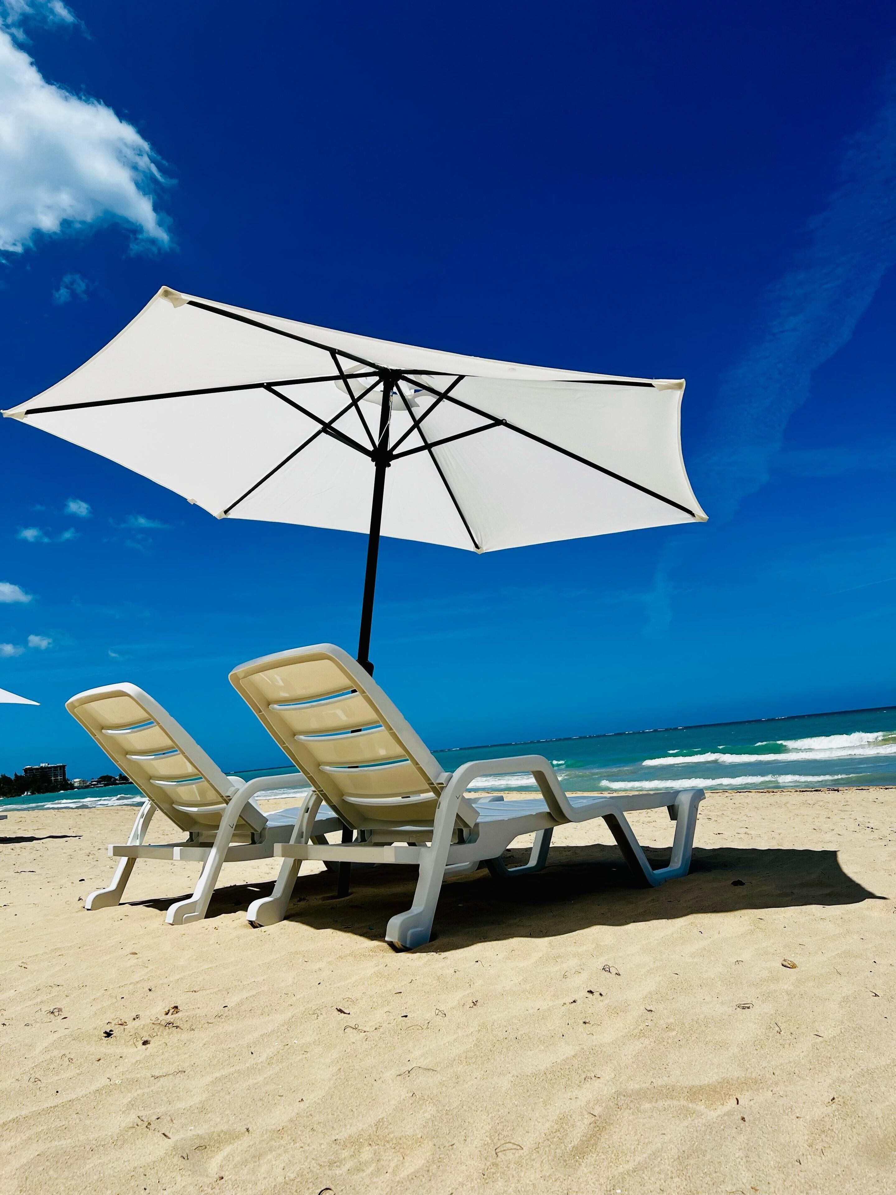 Two beach chairs under a white umbrella on a sandy beach with a clear blue sky and ocean in the background.