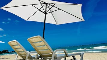 Two beach chairs under a white umbrella on a sandy beach with a clear blue sky and ocean in the background.