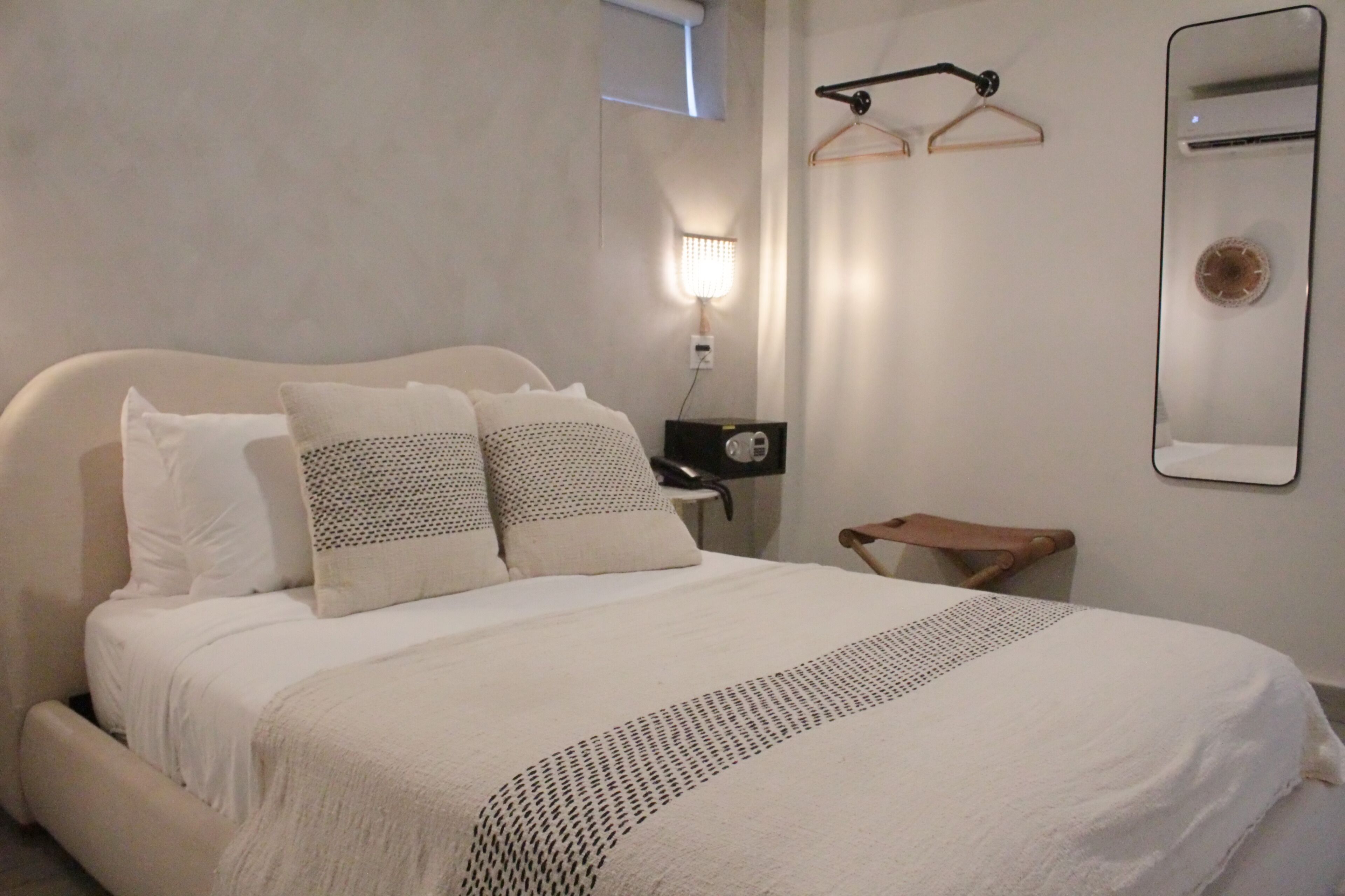 Una cama bien hecha con una colcha blanca y almohadas, una mesita de noche con una lámpara y un espejo fijado a la pared.