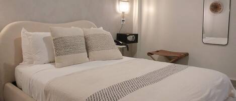 Una cama bien hecha con una colcha blanca y almohadas, una mesita de noche con una lámpara y un espejo fijado a la pared.