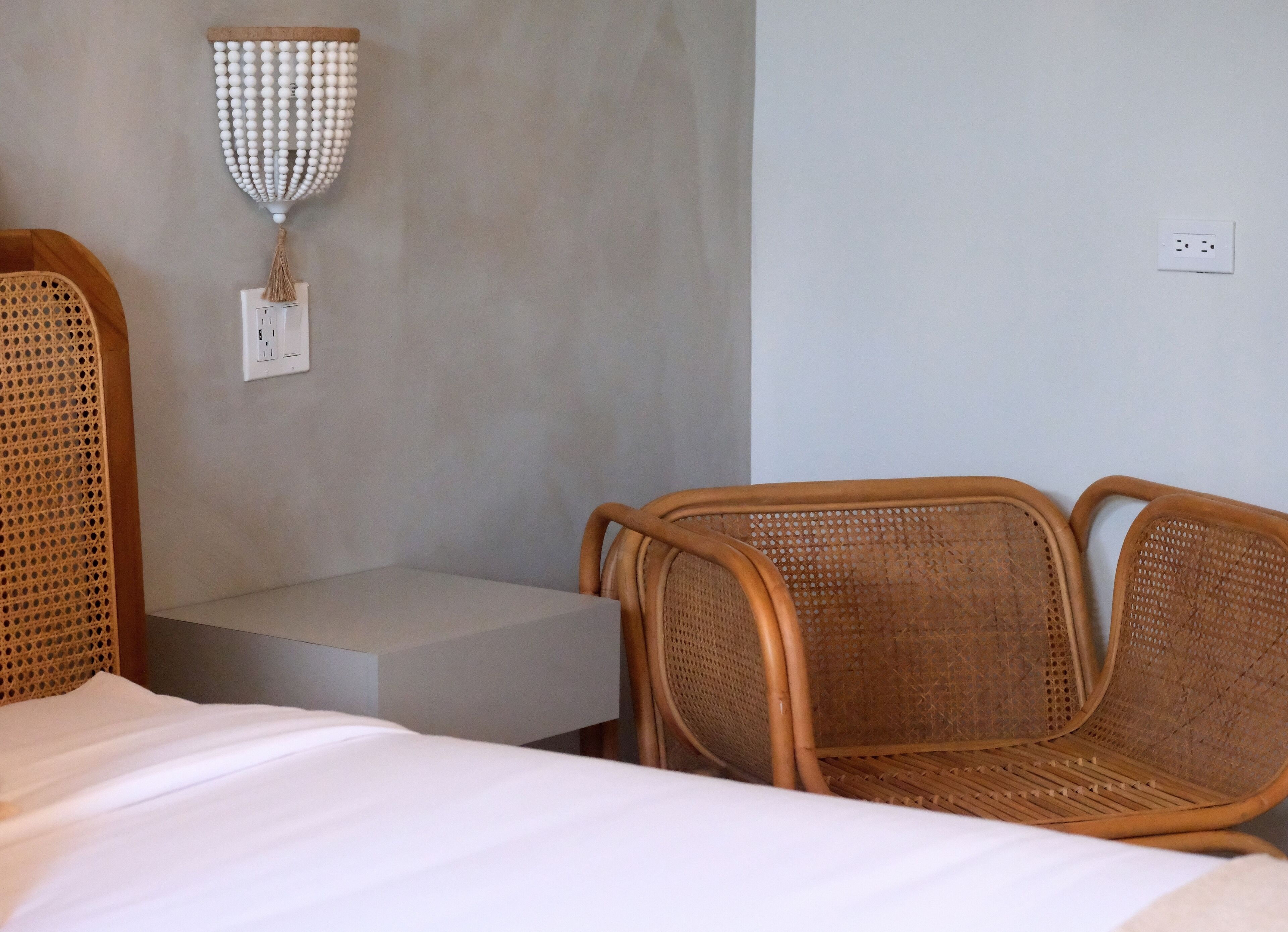 A hotel room with a bed, a small table, two wicker chairs, a wall-mounted light fixture, and a power outlet.