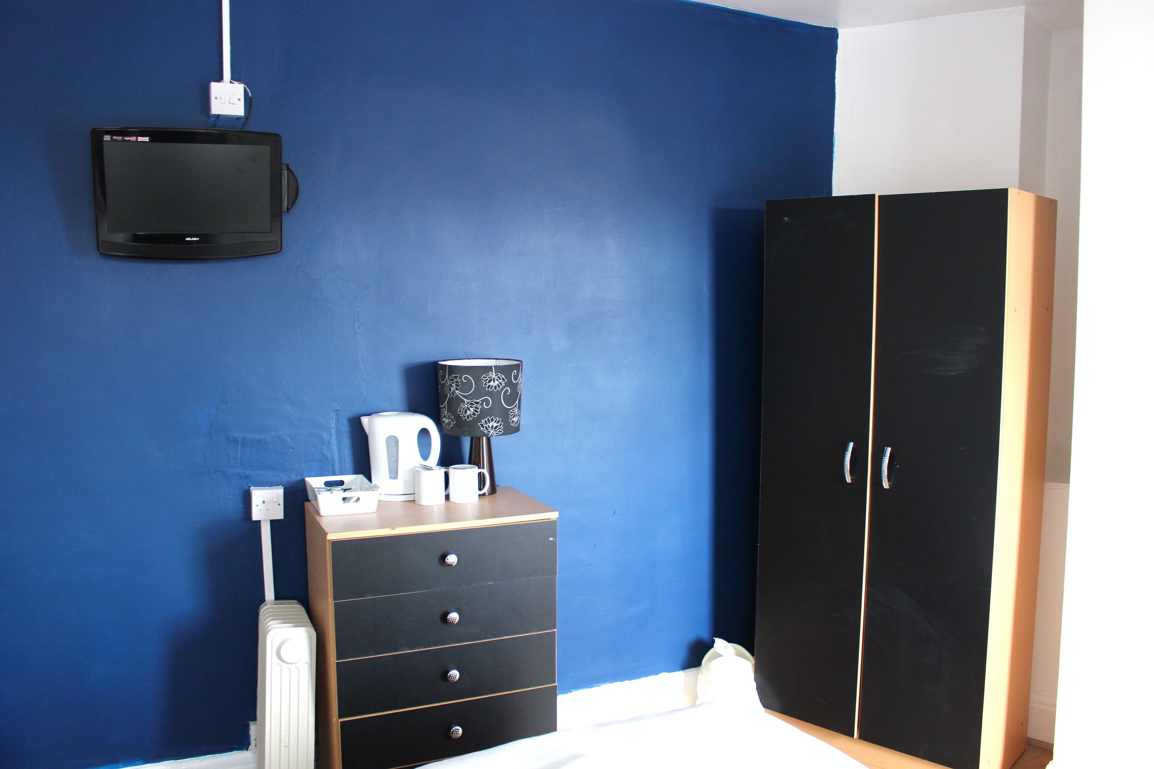 A room with a blue wall, a black wardrobe, a wooden dresser with drawers, a flat-screen TV mounted on the wall, and a white power outlet.