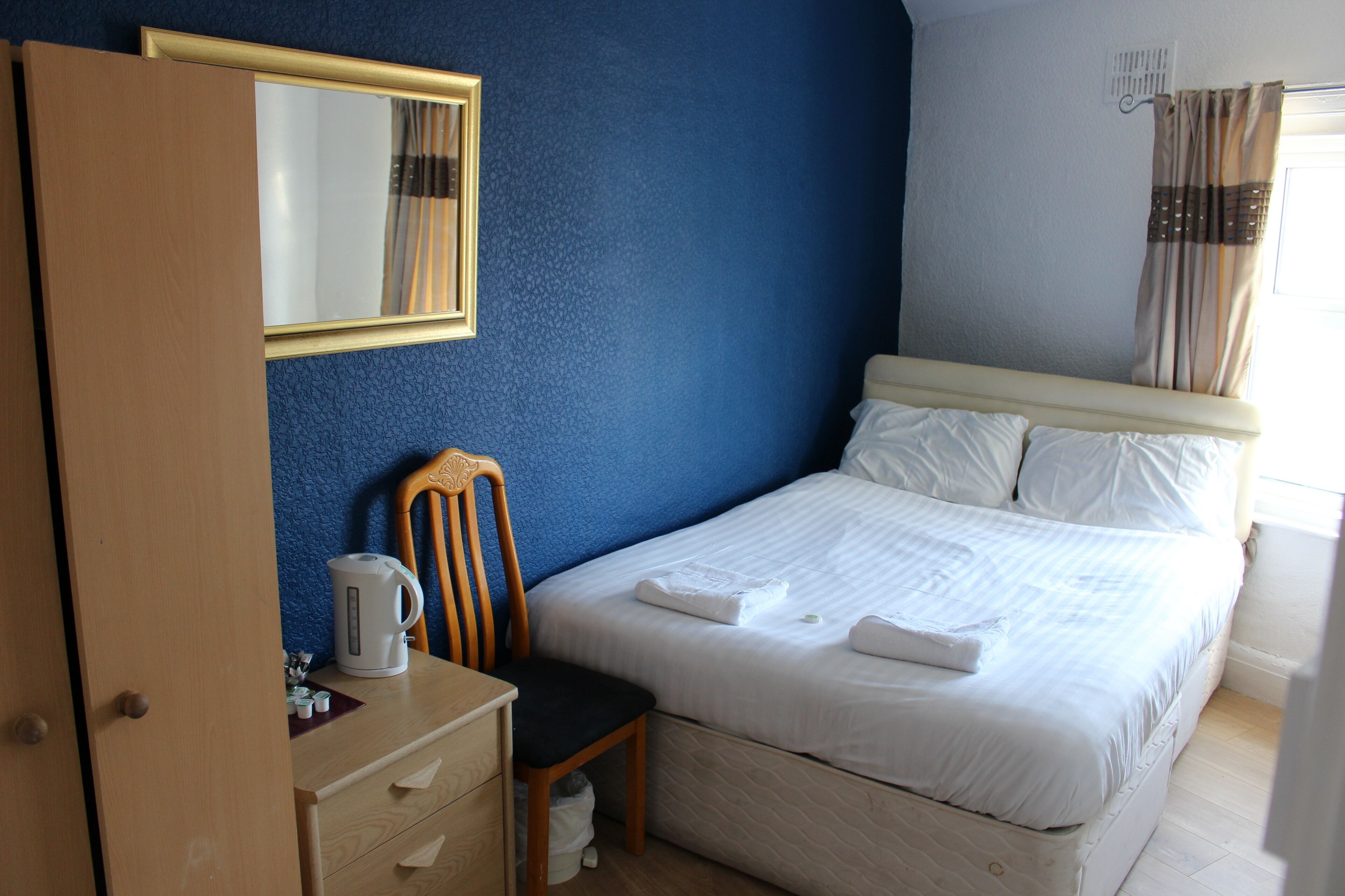 A hotel room with a bed, a chair, a desk with a kettle, and a mirror on the wall.