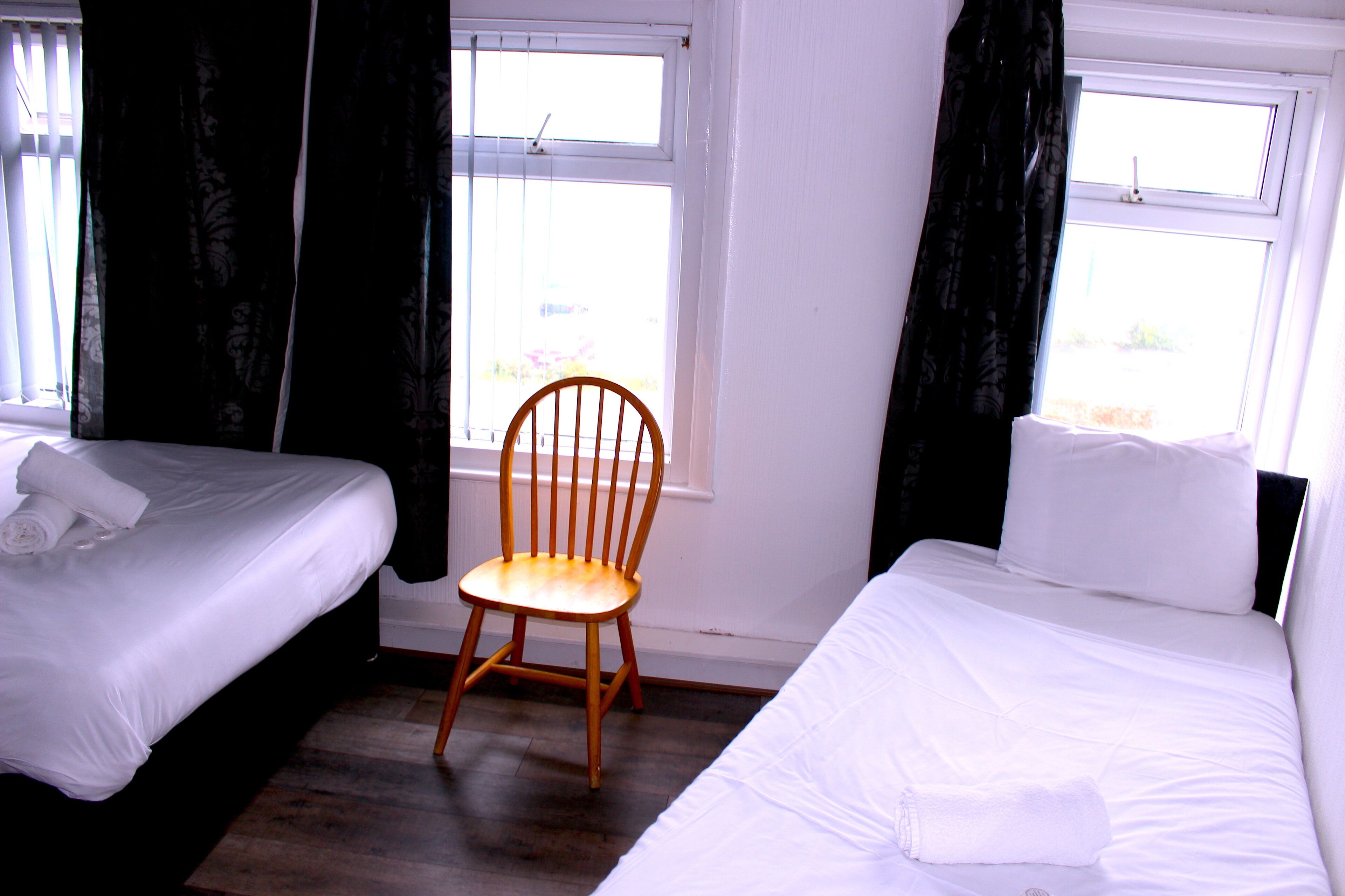 Una habitación de hotel con dos camas individuales, una silla de madera y un ventanal con cortinas.