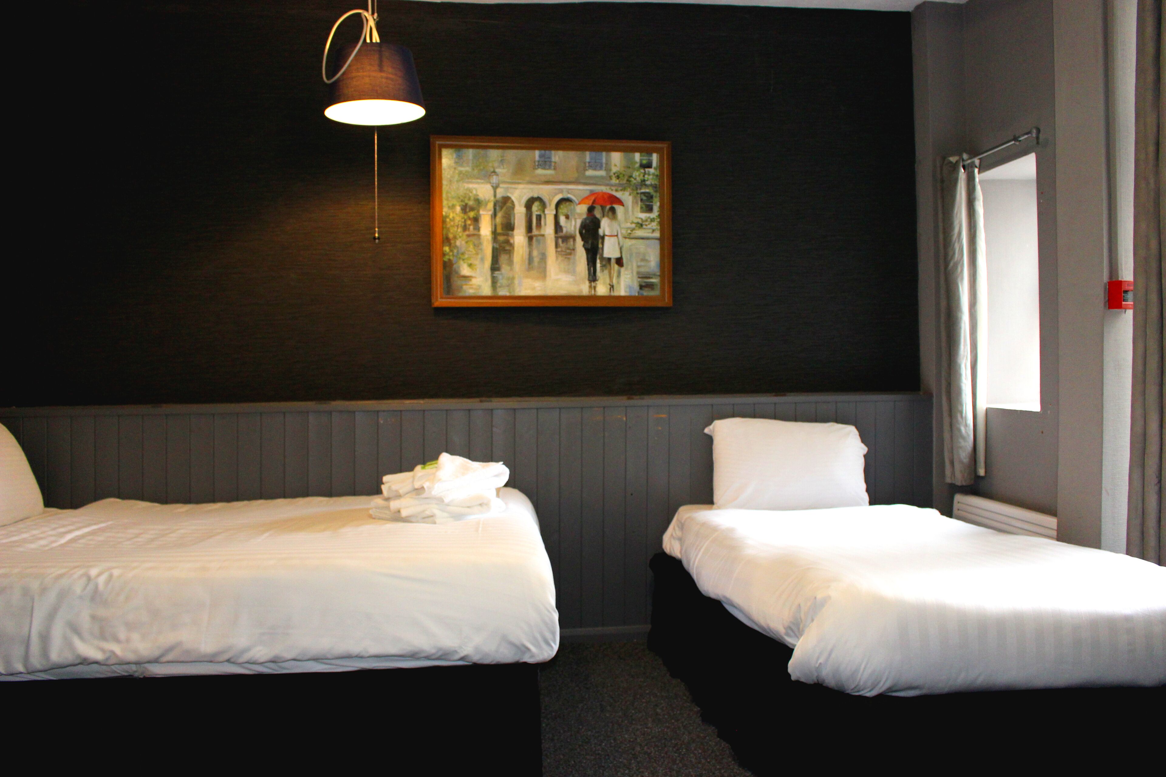 A hotel room with two beds, a painting of a couple on an umbrella, a window, and a lamp.