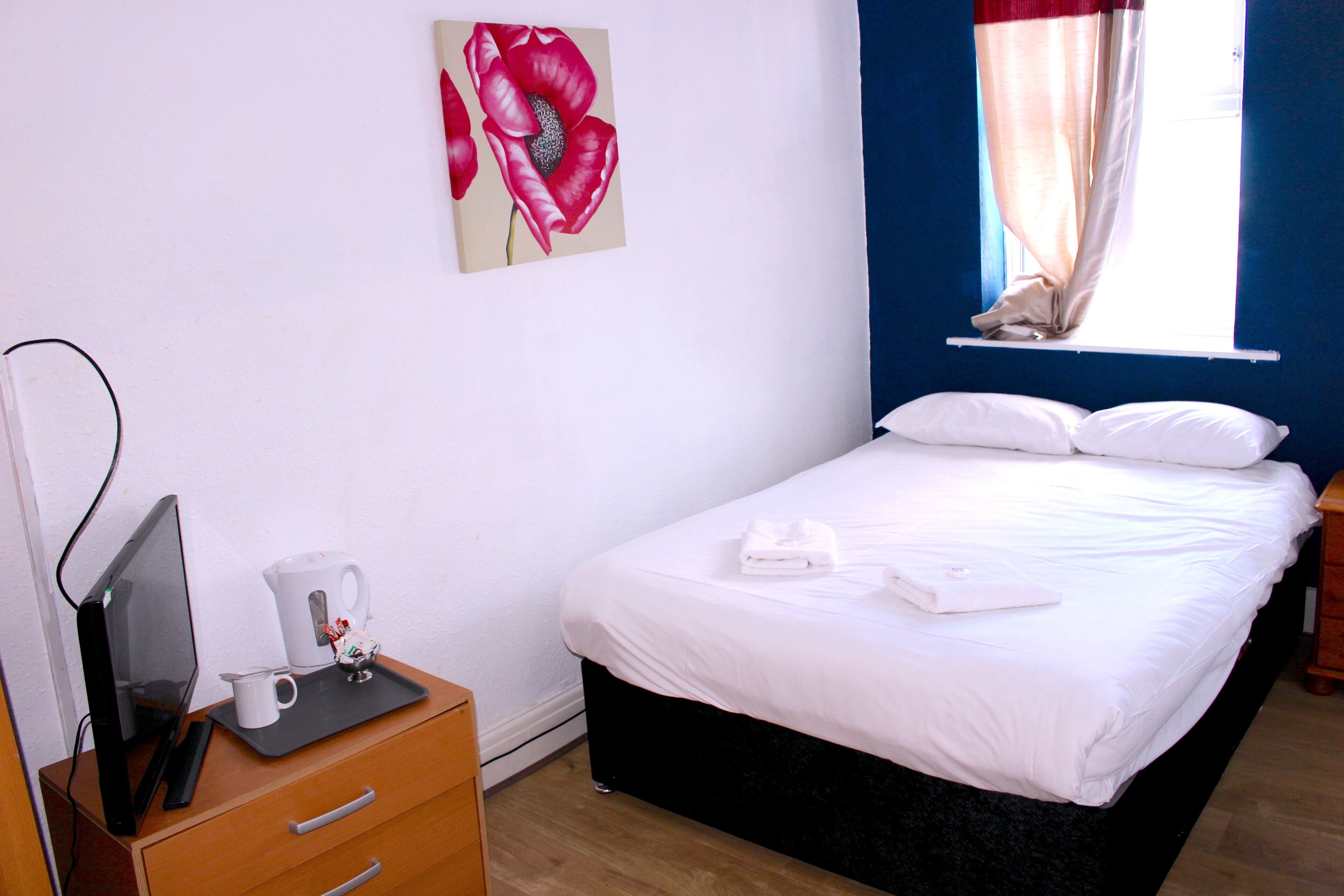 A neatly made bed with white linens, a bedside table with a coffee maker, and a painting of red flowers on the wall.
