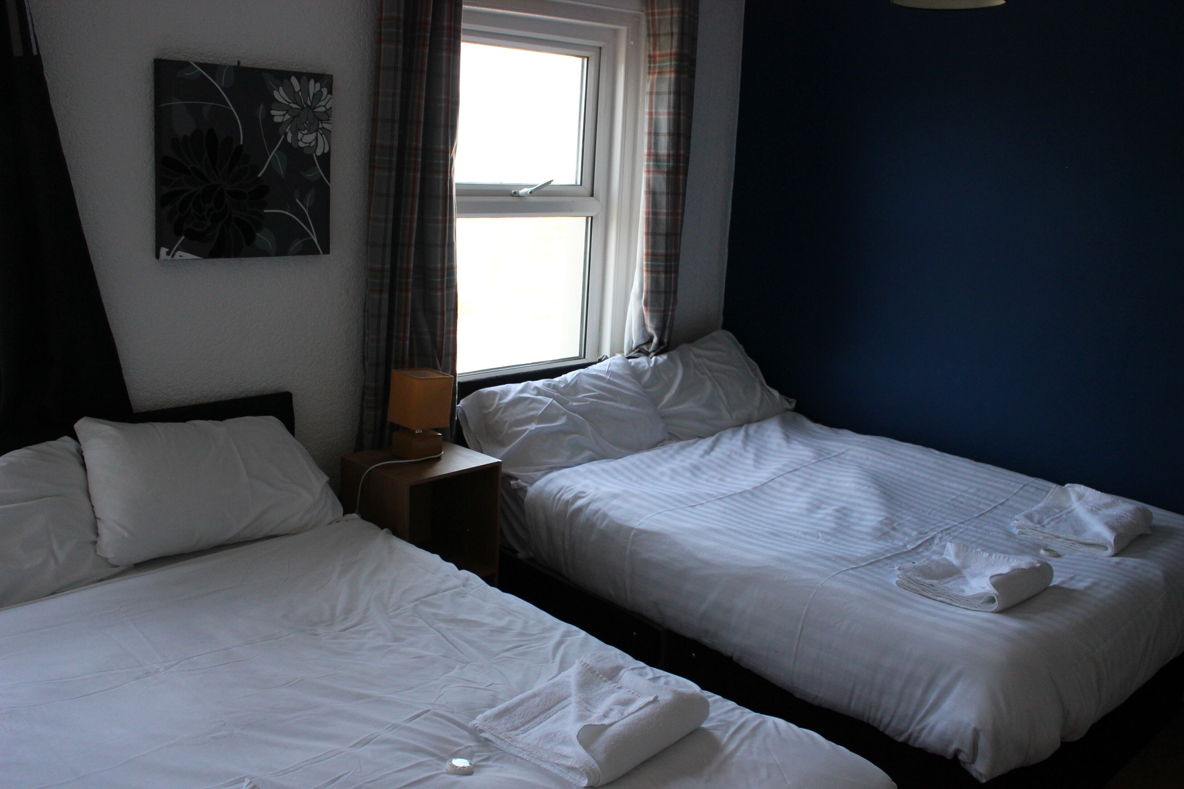 Quarto de hotel com duas camas de solteiro, uma mesa de cabeceira com abajur e janela com cortinas.