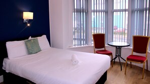A hotel room with a large bed, two red chairs, a small table, and a window with blinds. - Henson Hotel (Blackpool)