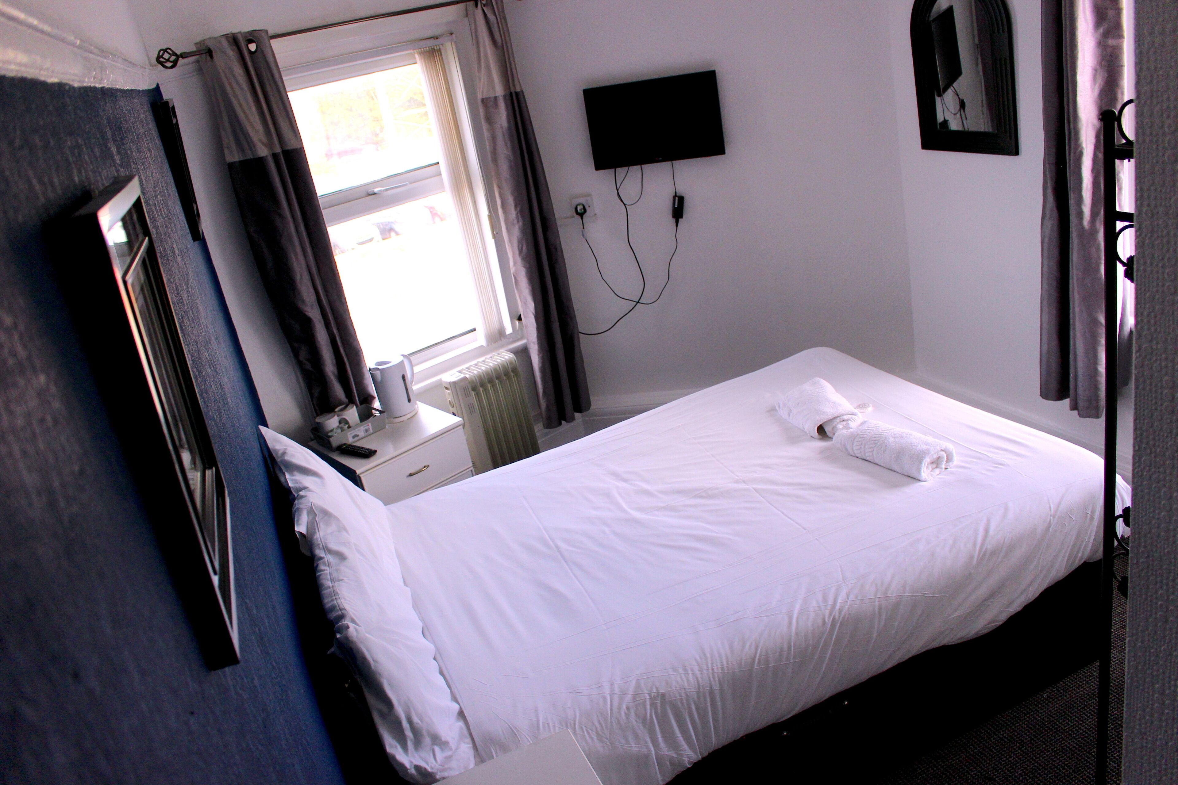 Ein Einzelbett mit weißer Bettwäsche, ein Nachttisch mit Lampe, ein Flachbildfernseher an der Wand und ein Fenster mit Vorhängen.