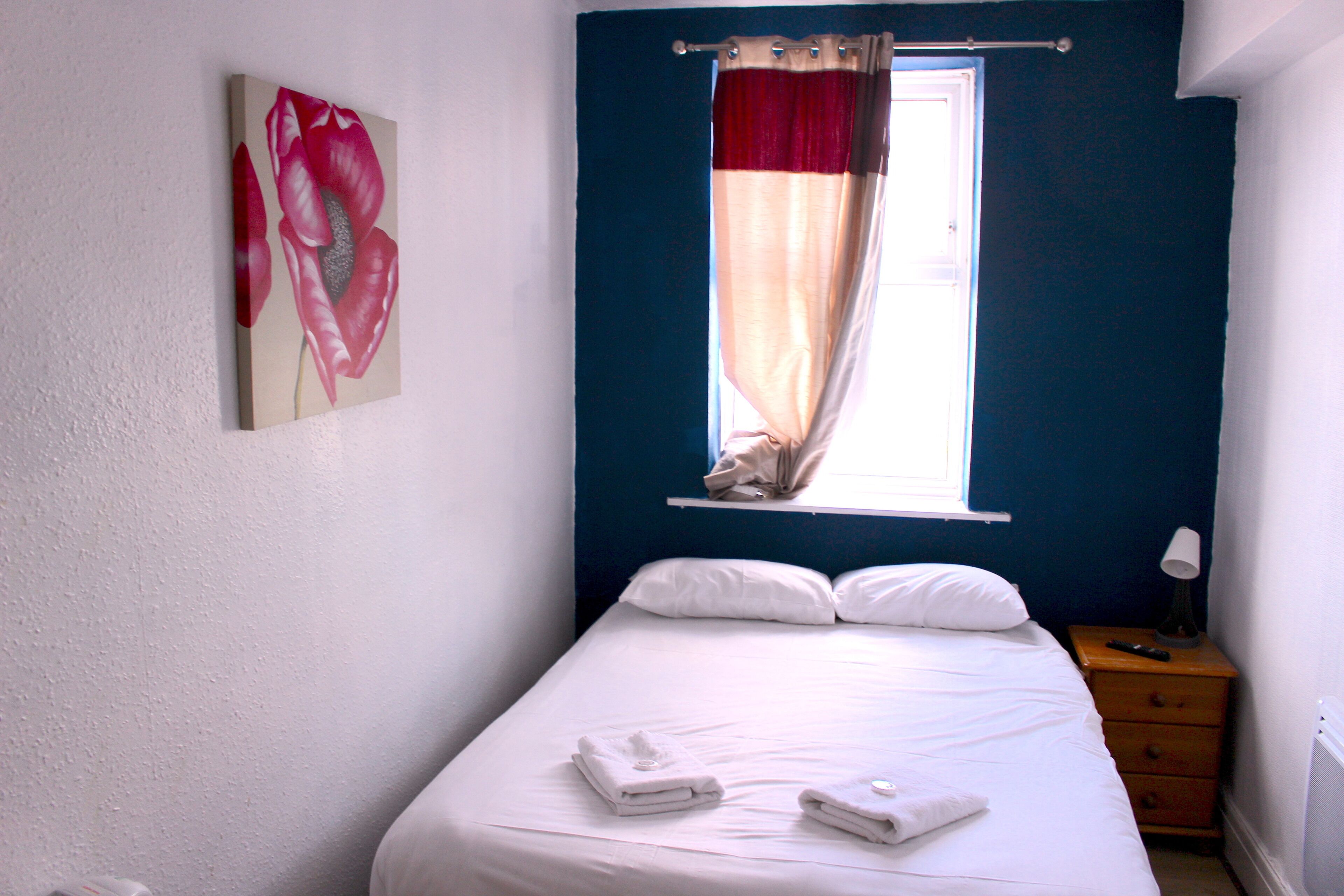 A bedroom with a bed, a window with curtains, a bedside table with a lamp, and a wall-mounted painting.