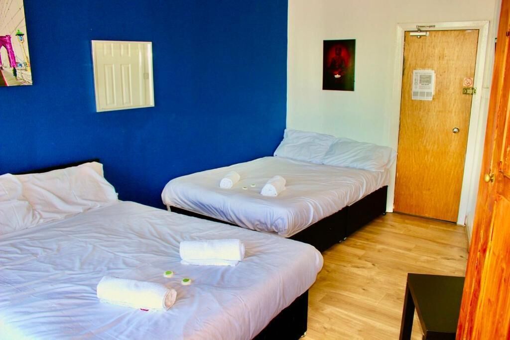 A room with two beds, a wooden door, a light switch, and a small table.
